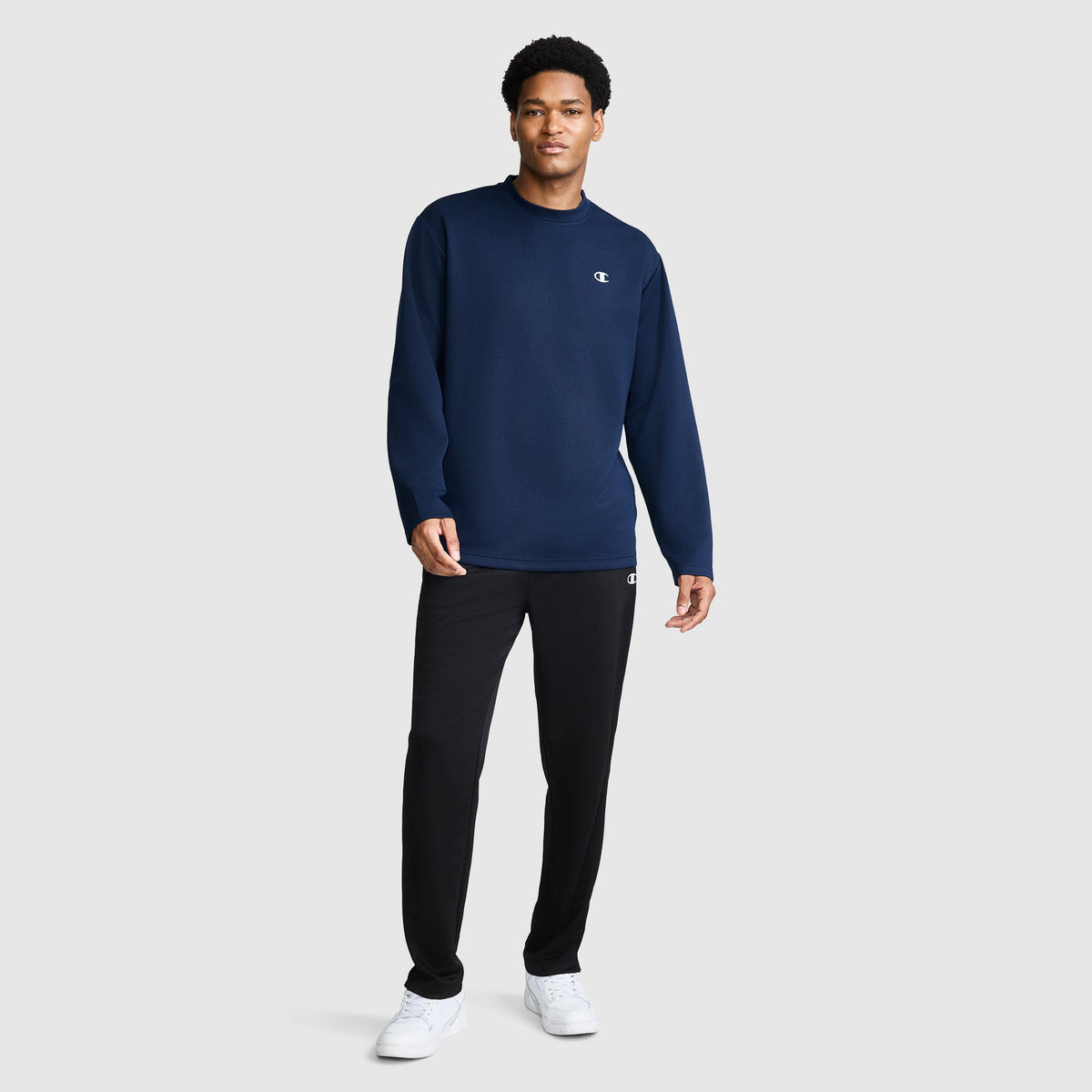 A man stands against a plain background wearing a navy blue Champion Sport Long Sleeve Crewneck, C Logo top, black pants, and white sneakers.