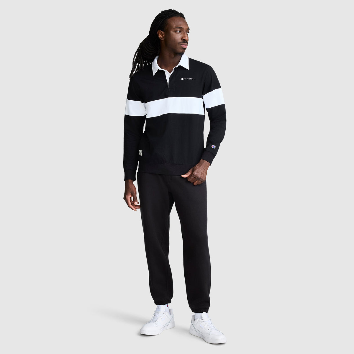 A man stands facing left, showcasing an athletic style in a black and white Champion Striped Rugby Shirt, paired with black pants and white sneakers, against a plain light gray background.