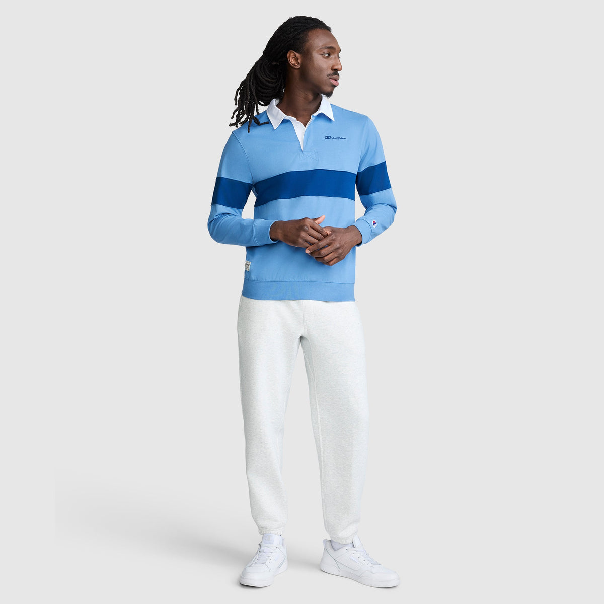 A man with long hair is seen wearing the Champion Striped Rugby Shirt in light blue with dark blue stripes, styled with white pants and sneakers for a laid-back athletic look against a simple light background.