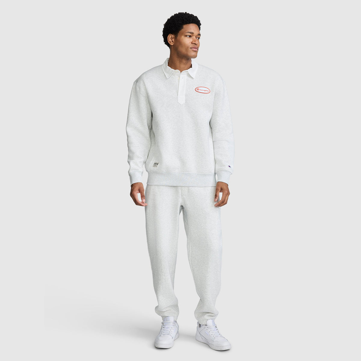 A person stands wearing a light gray Champion Fleece Rugby pullover and matching sweatpants with white sneakers. The Champion Fleece Rugby features a collar, buttons, and a red Champion logo on the chest. The background is plain and light.