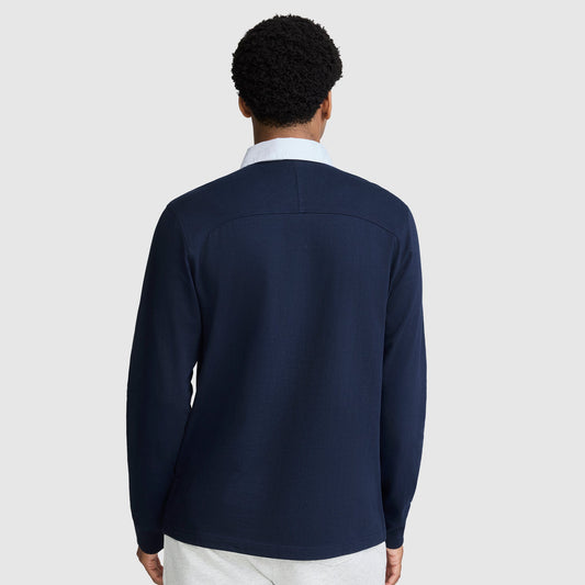 A person with short curly hair is shown from the back, wearing a Champion Long Sleeve Rugby Shirt with Twill Collar and Embroidered C Logo, layered over a white collared shirt and paired with light-colored pants, standing against a plain light gray background.