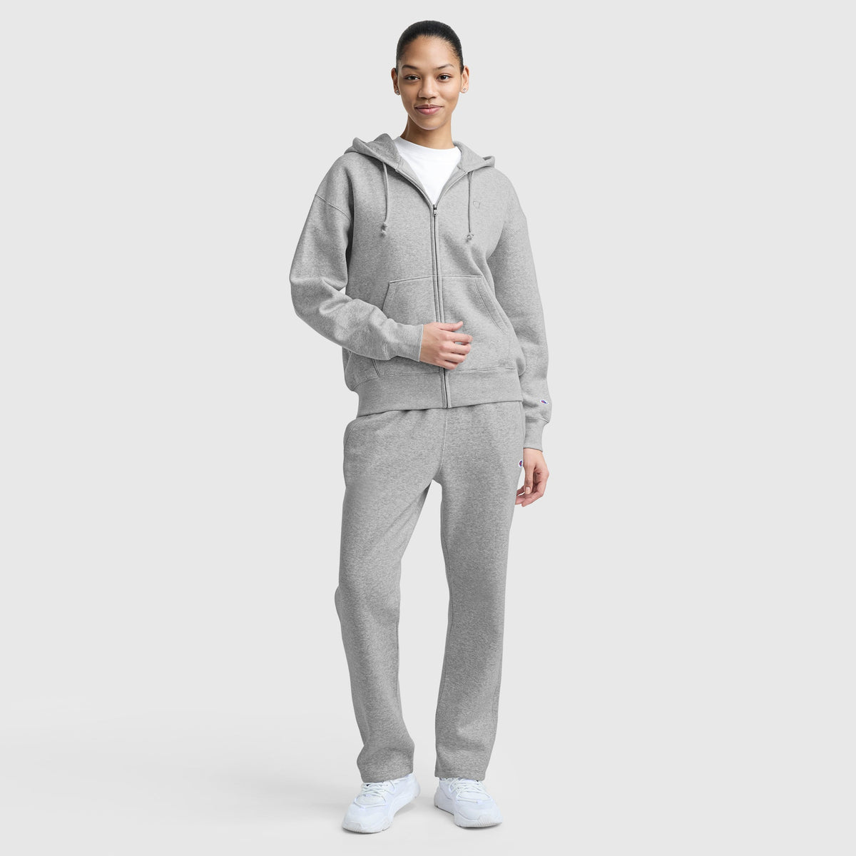 A person stands against a plain background, smiling softly with one hand on their jacket. They're wearing a Champion Powerblend Fleece Zip-Up Hoodie with C Logo in light gray, matching sweatpants, a white t-shirt, and white sneakers.