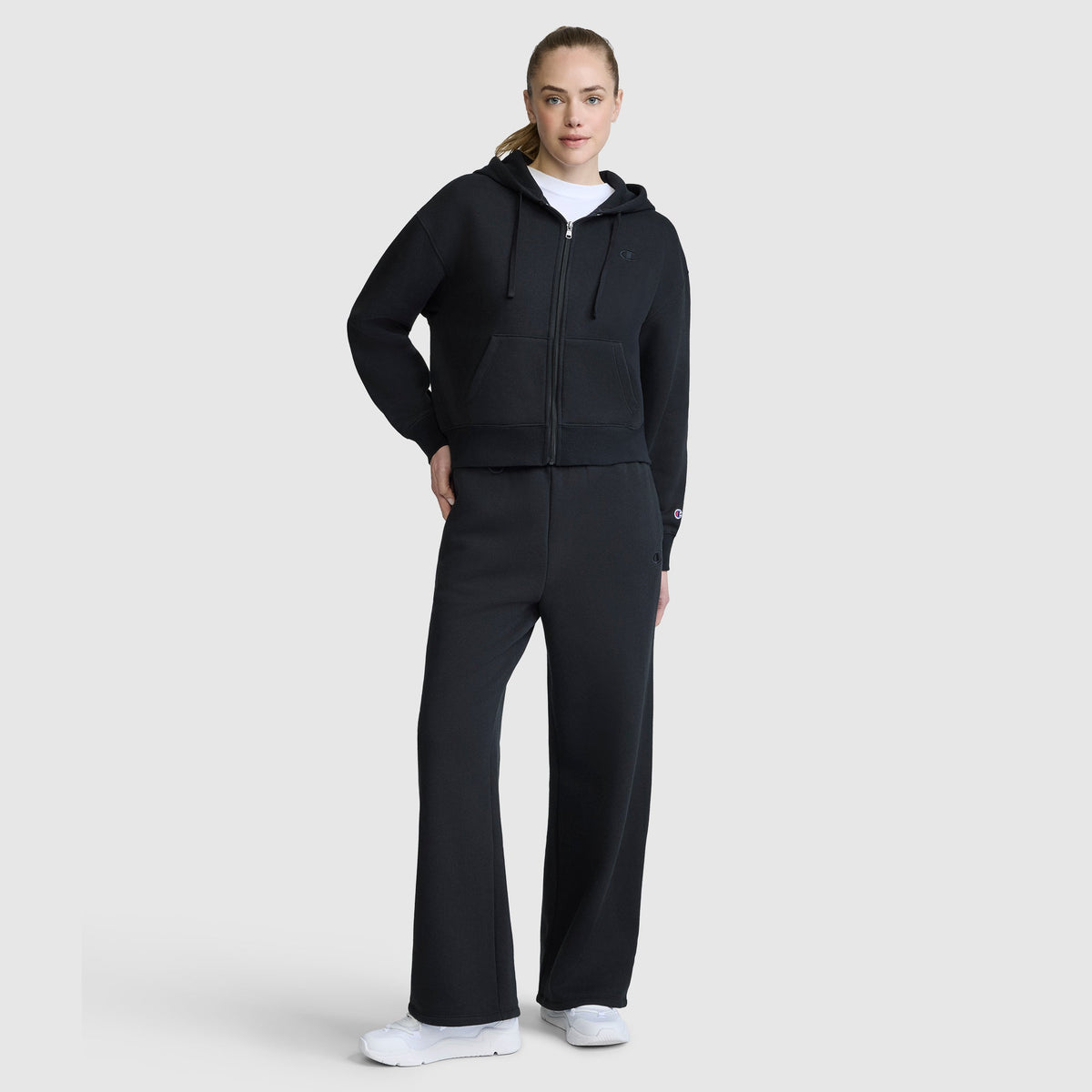 A woman poses against a plain light background, wearing a Champion Cropped Zip-Up Hoodie—an effortlessly stylish black fleece hoodie with a zip-up front—paired with matching wide-leg sweatpants, a white shirt, and white sneakers.