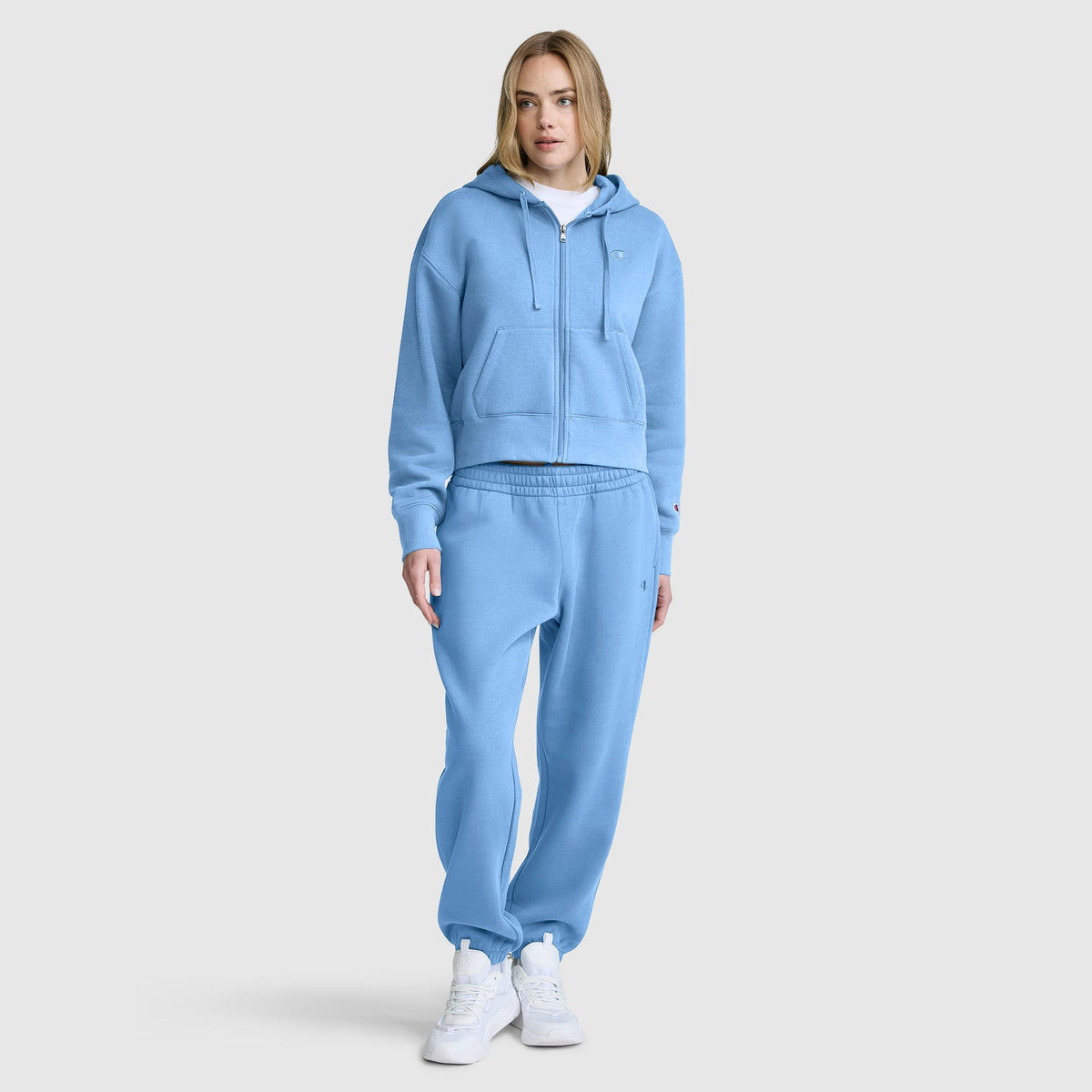 A blonde woman models a matching light blue Champion Cropped Zip-Up Hoodie and sweatpants set, featuring the cozy fleece hoodie paired with white sneakers against a plain gray background.