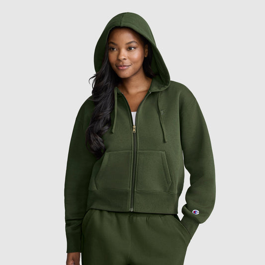 A woman wearing a dark green Champion Cropped Zip-Up Hoodie with the hood up and matching sweatpants stands facing forward, smiling slightly, against a plain light gray background.