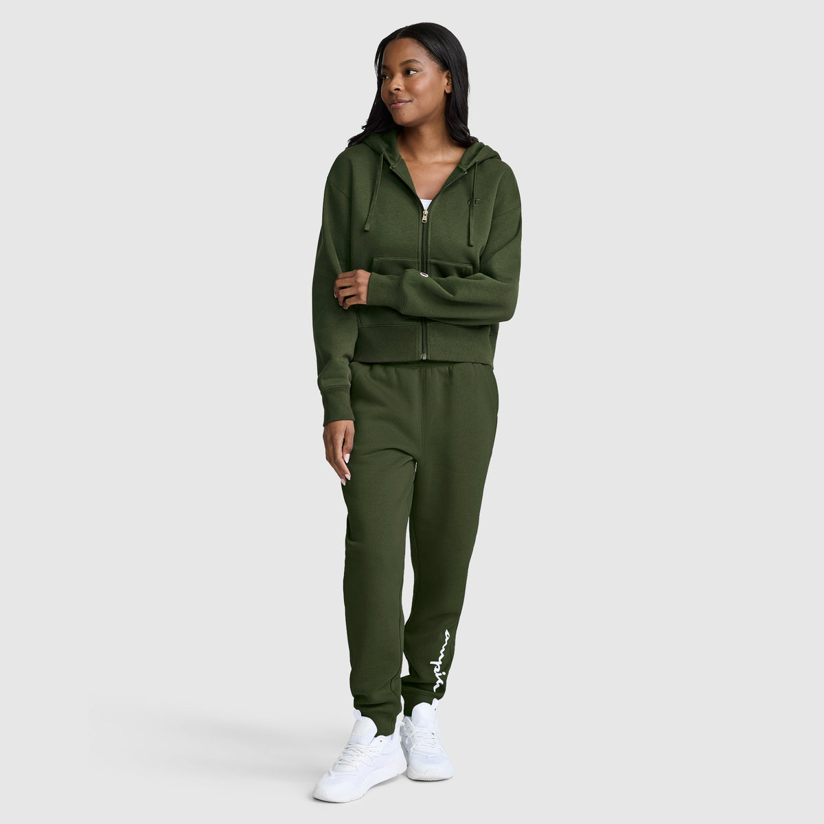 A woman wearing a dark green Champion Cropped Zip-Up Hoodie with matching joggers and white sneakers stands against a plain light background, looking to the side.