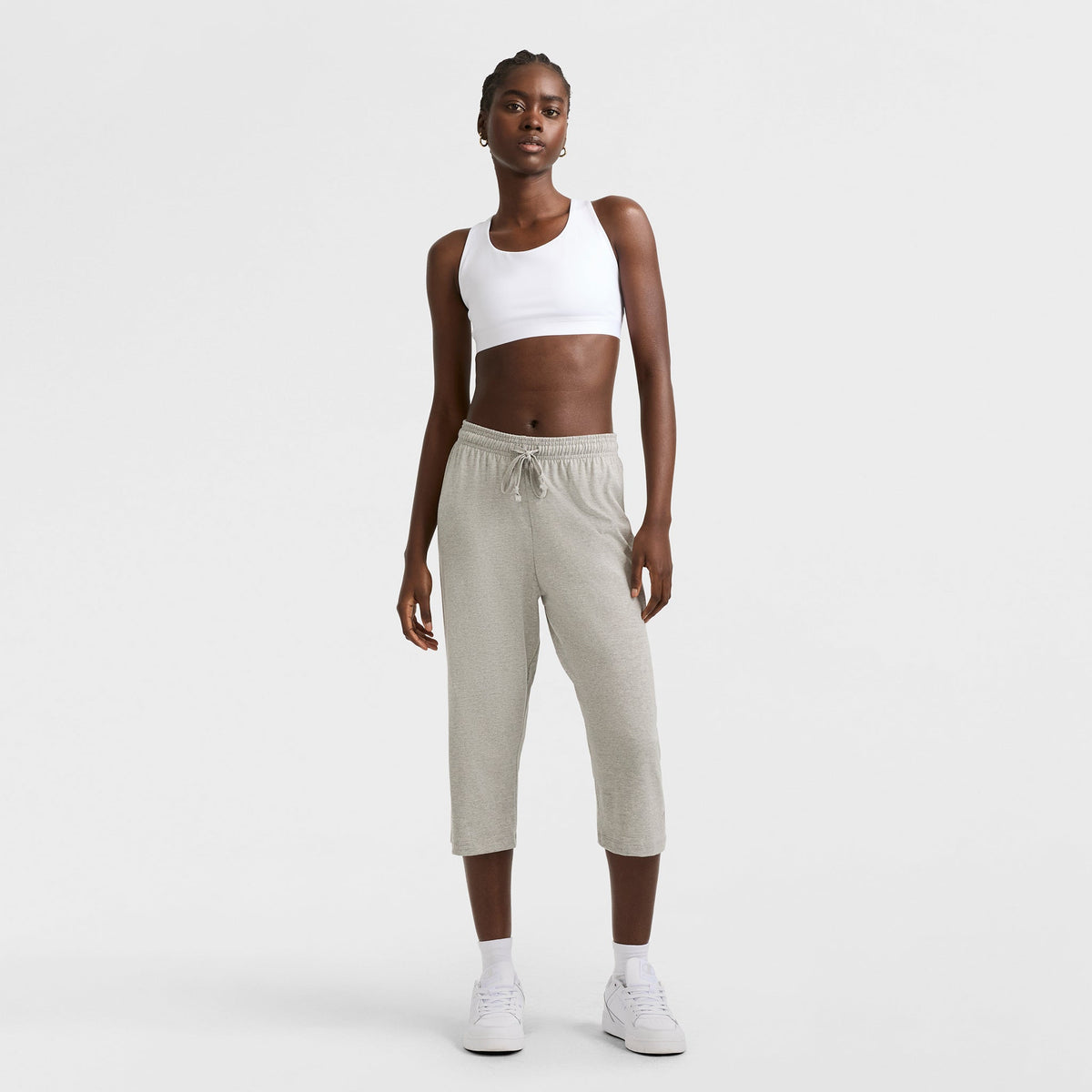 Against a white background, a woman models Champion's Lightweight Lounge Capri Pants, C Logo, in light gray with a white sports bra and sneakers. She stands relaxed and confident, looking directly at the camera.