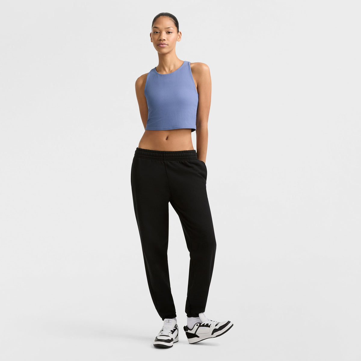 A woman stands against a plain white background, wearing Champion's Powerblend Oversized Sweatpants with a C Logo in black, paired with a sleeveless blue crop top and white sneakers. She has her hands behind her back and looks forward neutrally.
