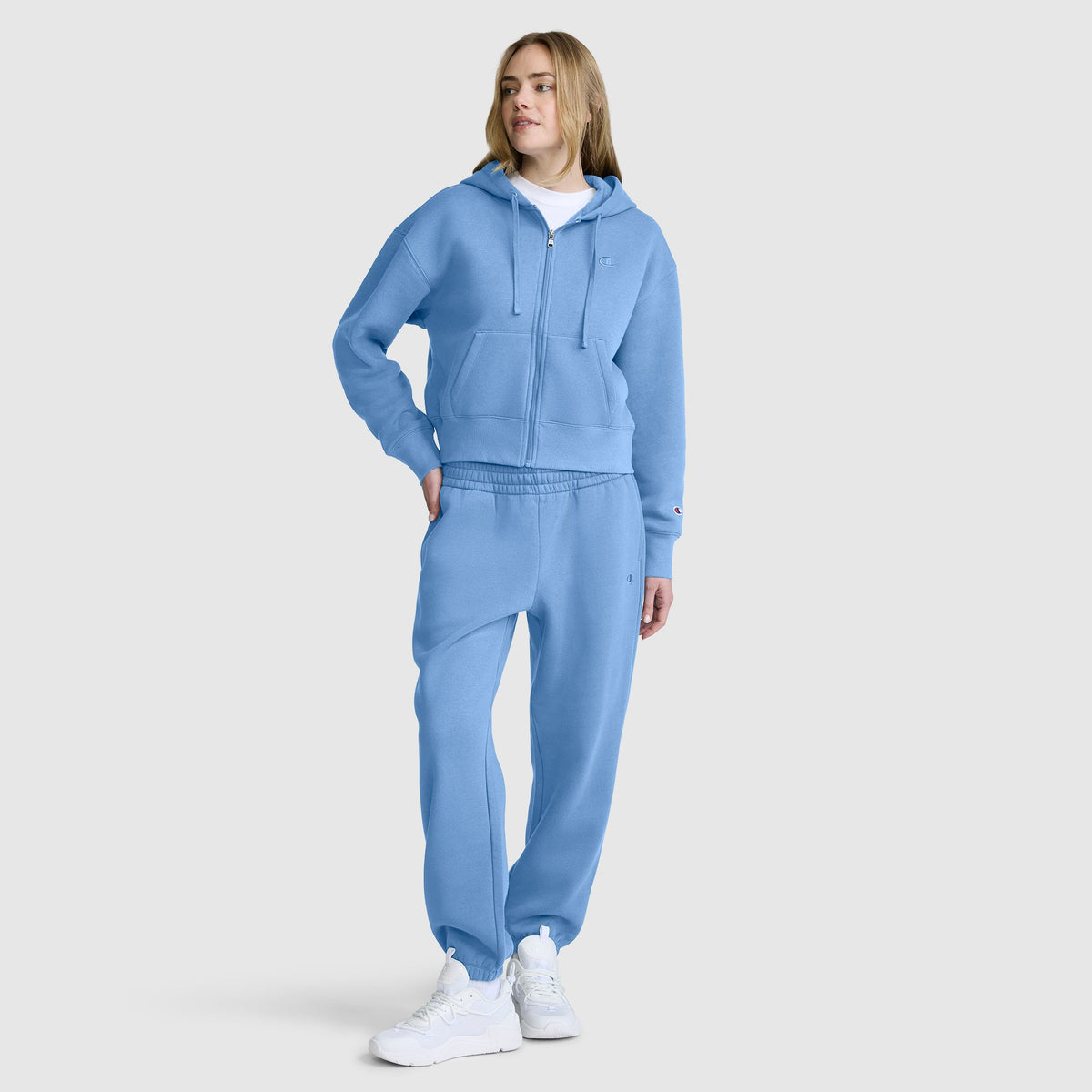 A person wearing a light blue hooded sweatshirt and Champion Powerblend Oversized Sweatpants with the C logo stands with one hand in their pocket. They also wear white sneakers and a white t-shirt, posing against a plain light gray background.