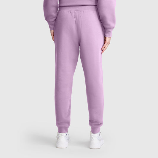 A person stands facing away, wearing Champion Powerblend Joggers with a C Logo in light purple matched with a coordinating sweatshirt and white sneakers. The background is plain and light-colored.