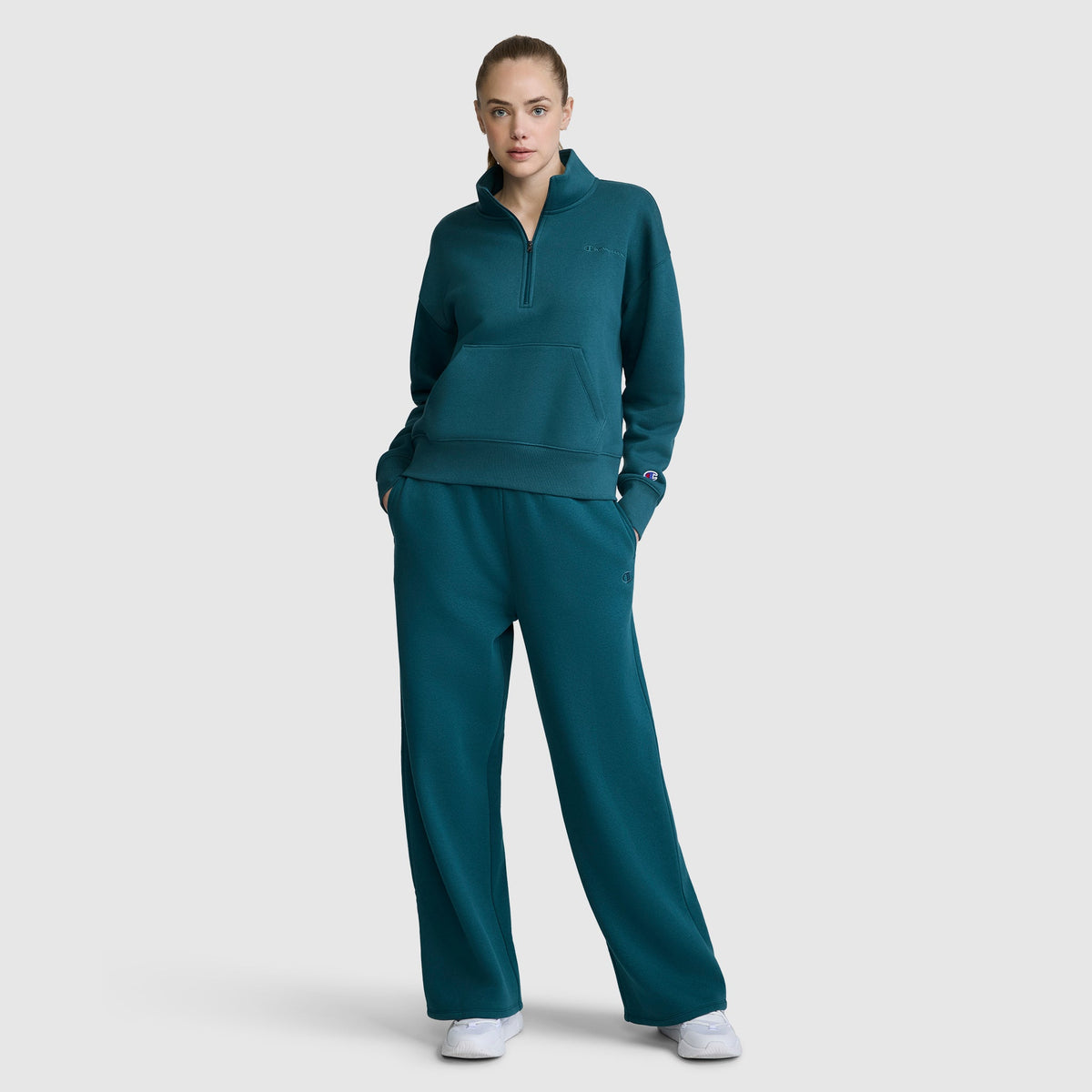 A person stands facing forward with hands in their pockets, wearing Champion’s Wide Leg Fleece Sweatpants with the C Logo in teal, paired with a matching pullover sweatshirt and white sneakers, against a plain light gray background.