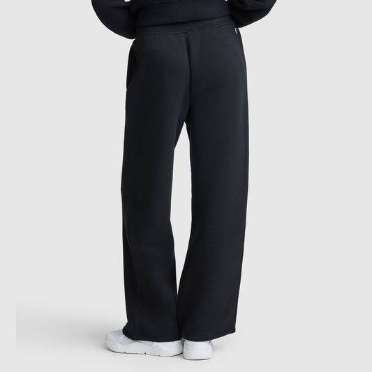 A person is seen from behind, standing against a light gray background, wearing Champion Wide Leg Fleece Sweatpants, C Logo in black paired with white sneakers. The full length of the women's sweatpants is visible in the photo.