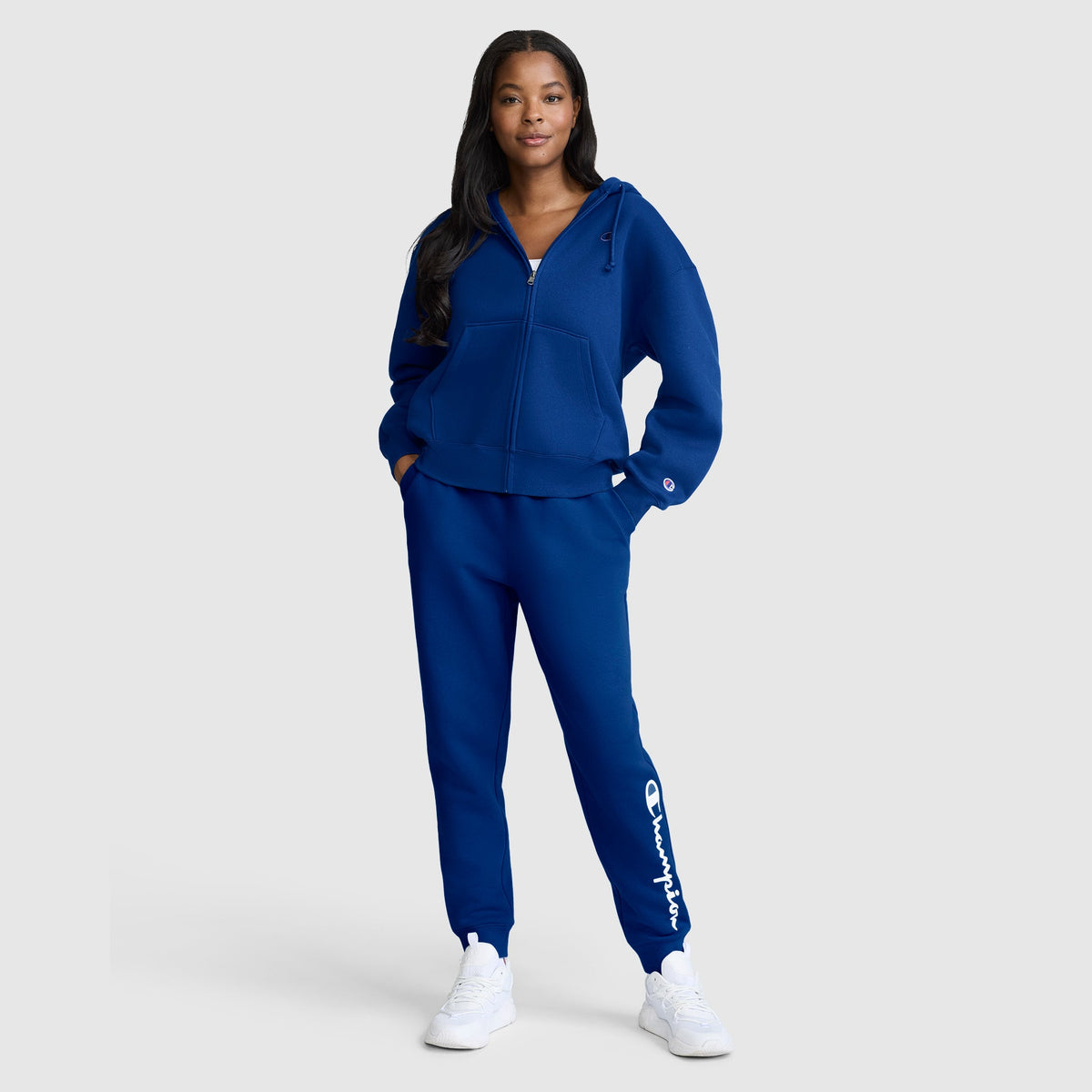 A woman stands smiling with her hands in the pockets of her matching blue Champion Powerblend Joggers with Classic Script Logo and a zip-up hoodie, paired with white sneakers, against a plain light background.