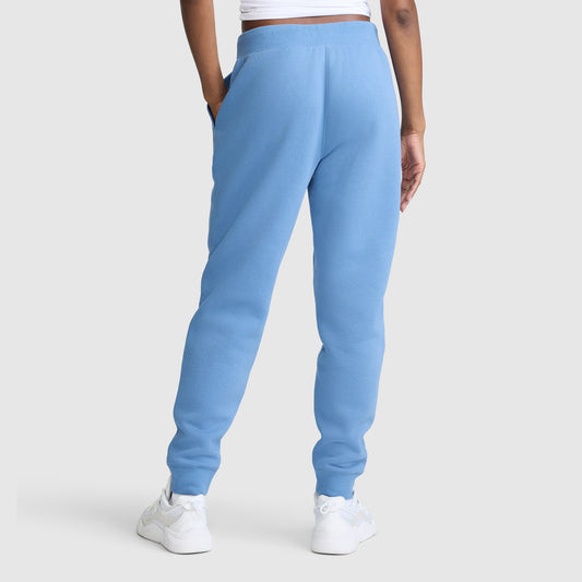 A person is shown from the back against a plain light gray background, wearing Champion Powerblend Joggers with the Classic Script Logo in light blue, paired with a white cropped top and white sneakers.