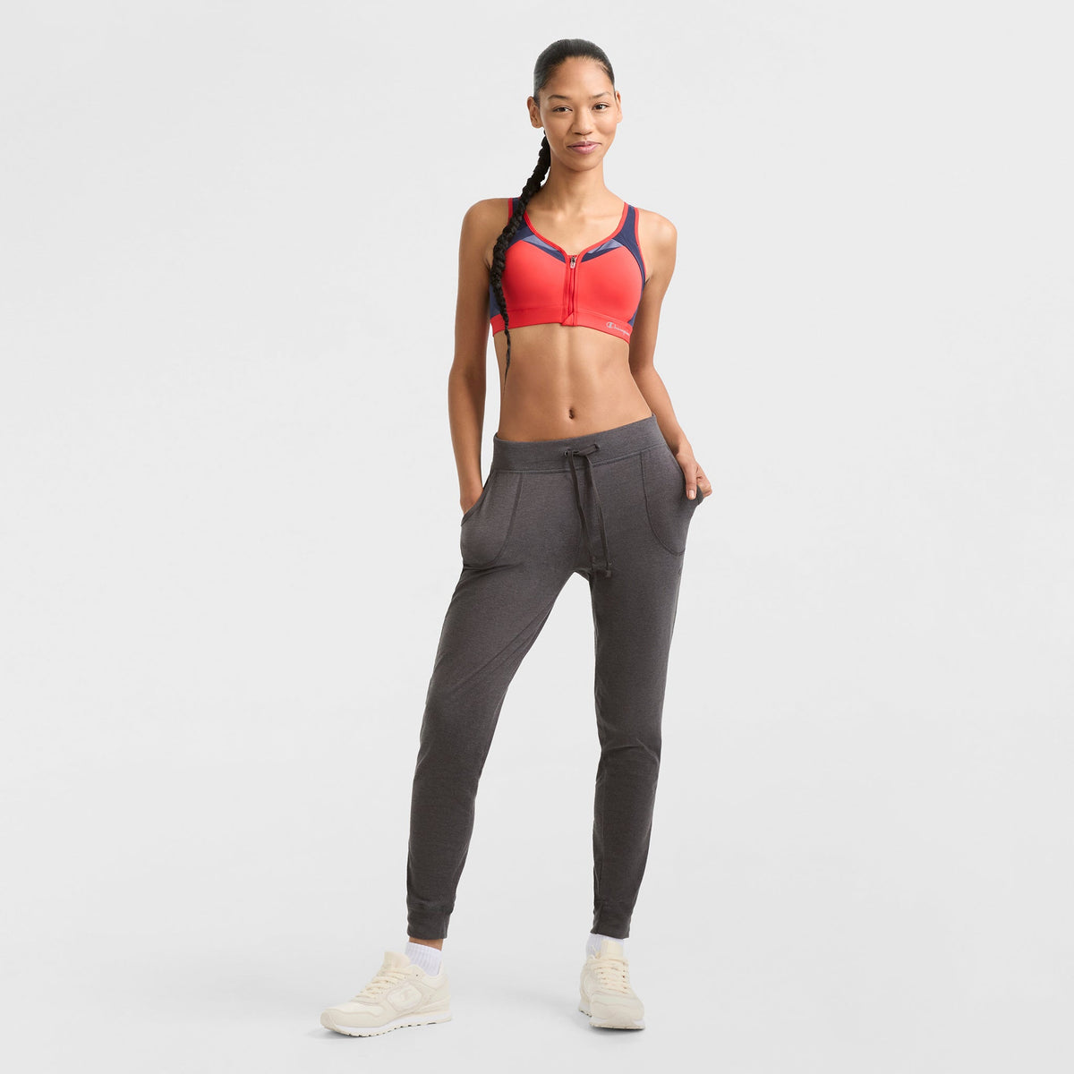 A woman stands confidently in a red sports bra, Champion Lightweight Lounge Joggers in dark gray with pockets, and white sneakers against a plain white background.