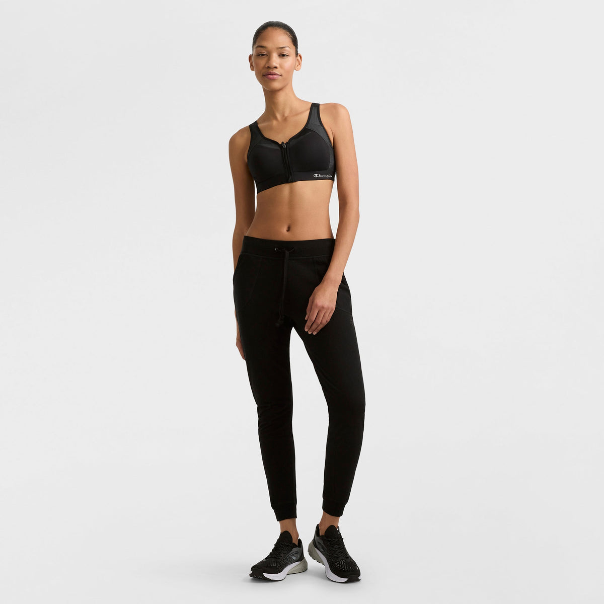 A woman stands against a white background, wearing Champion Lightweight Lounge Joggers, a black sports bra, and black sneakers. She poses casually with one hand at her side and a subtle smile.