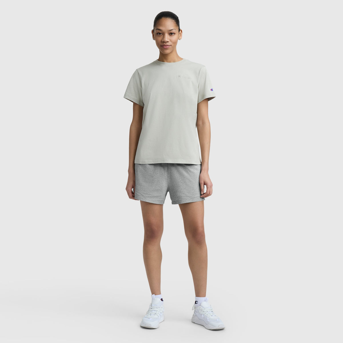 A person stands facing forward, wearing a light gray t-shirt, Champion Practice Shorts in gray, and white sneakers against a plain white background.