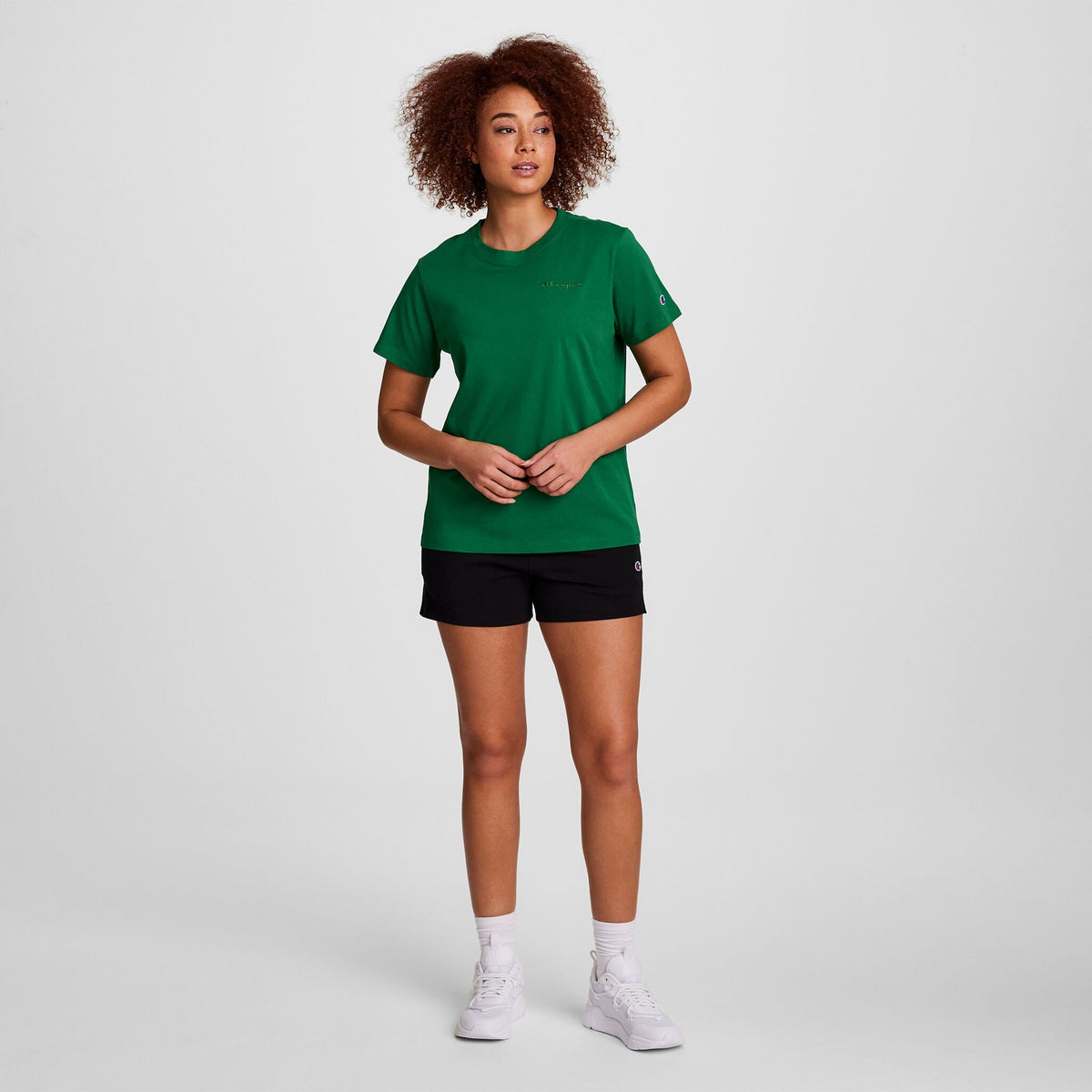 A person with curly hair stands against a white background, facing forward with hands loosely clasped, wearing a green t-shirt and Champion Practice Shorts for women, paired with white socks and white sneakers.