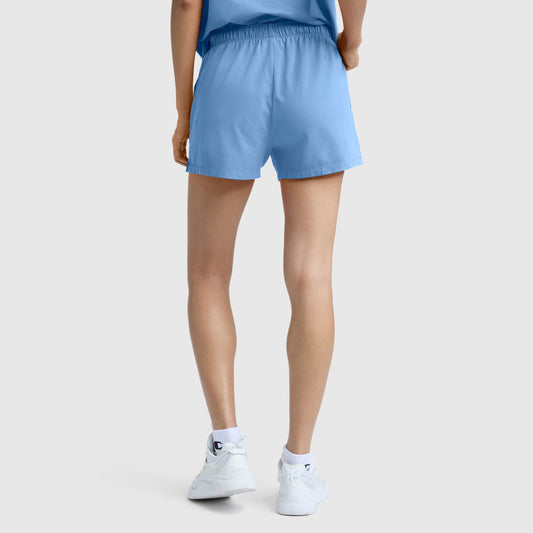 A person stands with their back to the camera, wearing a light blue T-shirt, Champion Practice Shorts in a matching light blue shade, and white sneakers against a plain background.