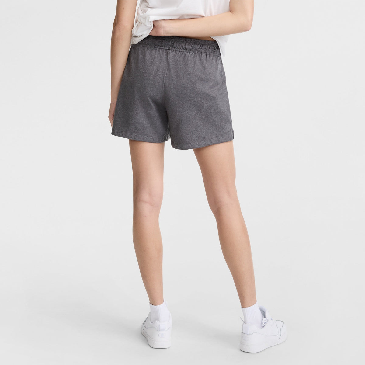 A person stands with their back to the camera, wearing a white t-shirt, Champion 4" Lightweight Lounge Shorts with C Logo in gray, white socks, and white sneakers against a plain white background.