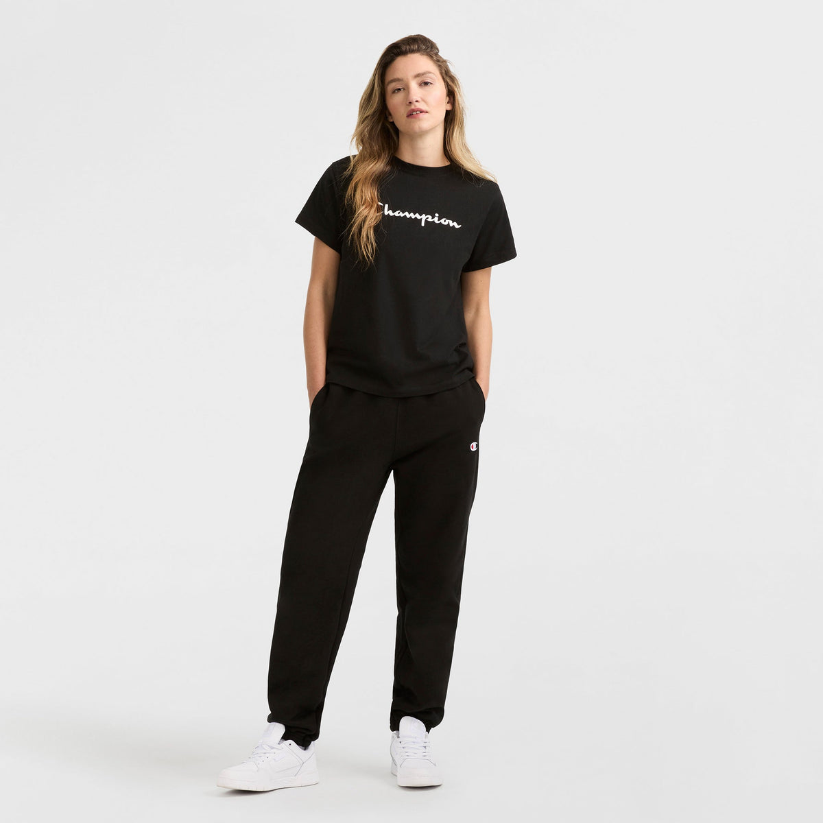 A person with long, wavy hair stands against a plain white background wearing the Champion Classic T-Shirt with script logo, black sweatpants, and white sneakers. Their hands are in their pockets.