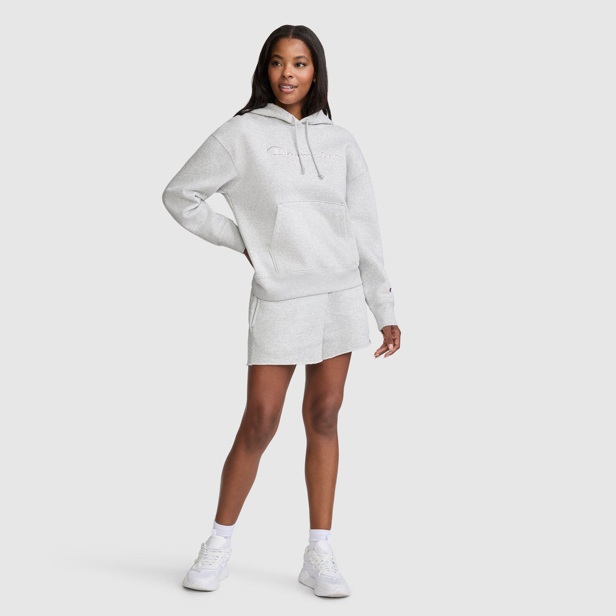 A woman stands in a Champion Tonal Script Freshman Powerblend Hoodie and matching shorts with white socks and sneakers, smiling with one hand on her hip against a plain light background.