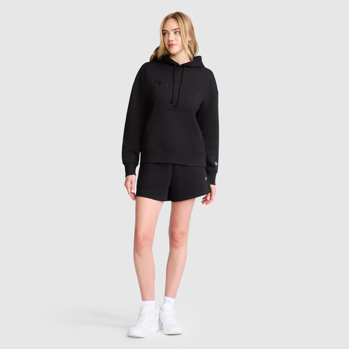 A woman stands against a plain background in a black Champion Tonal Script Freshman Powerblend Hoodie, paired with black shorts, white socks, and sneakers. Her light hair frames her face as she gazes aside, one hand at her side and the other on her thigh.