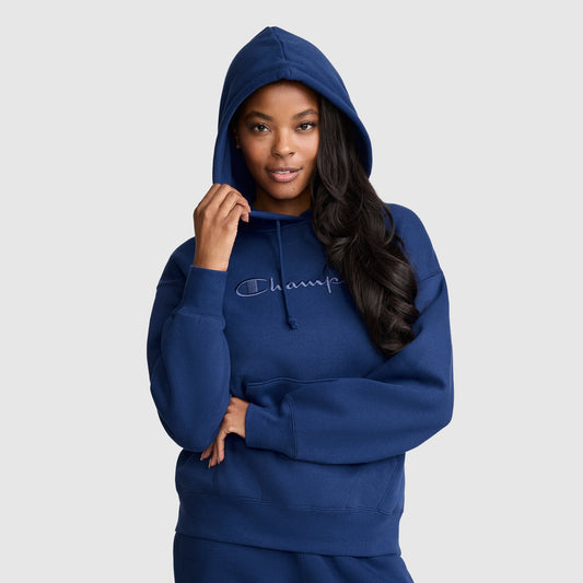 A woman wearing a blue Champion Tonal Script Freshman Powerblend Hoodie, crafted from soft midweight fleece, poses with one hand holding the hood and her other arm crossed at her waist against a plain light gray background.