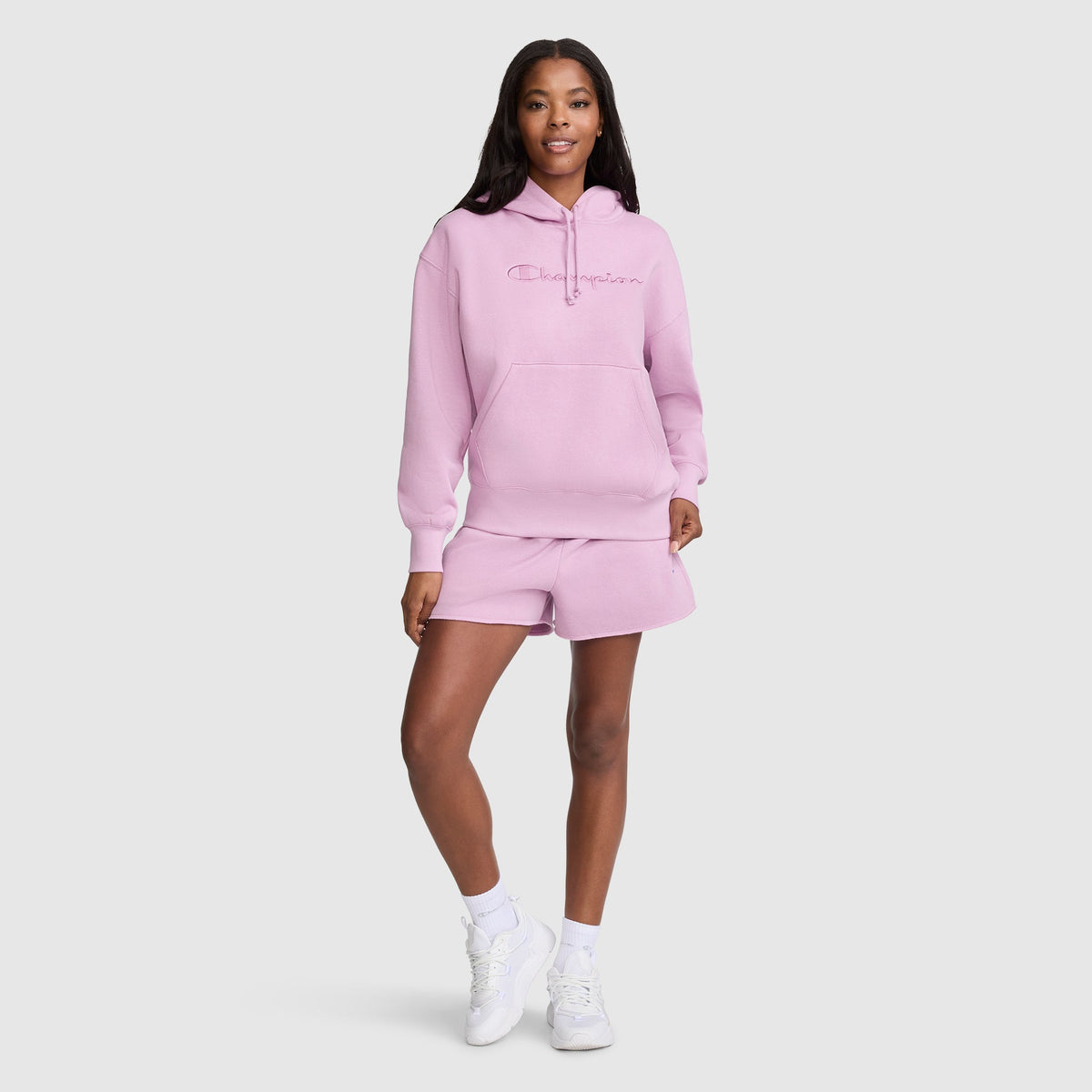 A woman stands smiling in a light pink Champion Tonal Script Freshman Powerblend Hoodie and matching shorts with white socks and sneakers, posing against a plain white background.