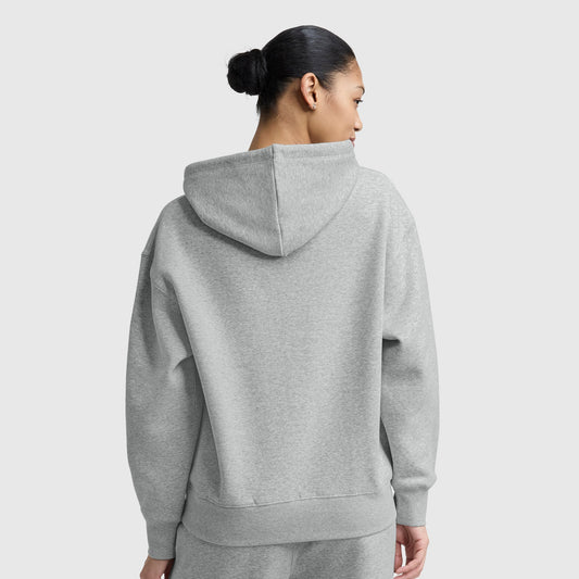 A woman with dark hair in a bun, seen from behind, wears the Champion Small C Freshman Powerblend Hoodie and matching sweatpants in light gray midweight fleece against a plain light gray background.