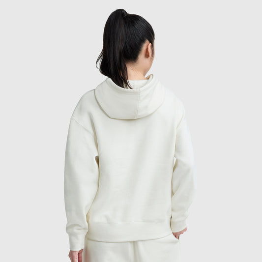 A person with long dark hair in a ponytail is seen from behind, wearing an off-white Champion Small C Freshman Powerblend Hoodie and matching pants against a plain light gray background.