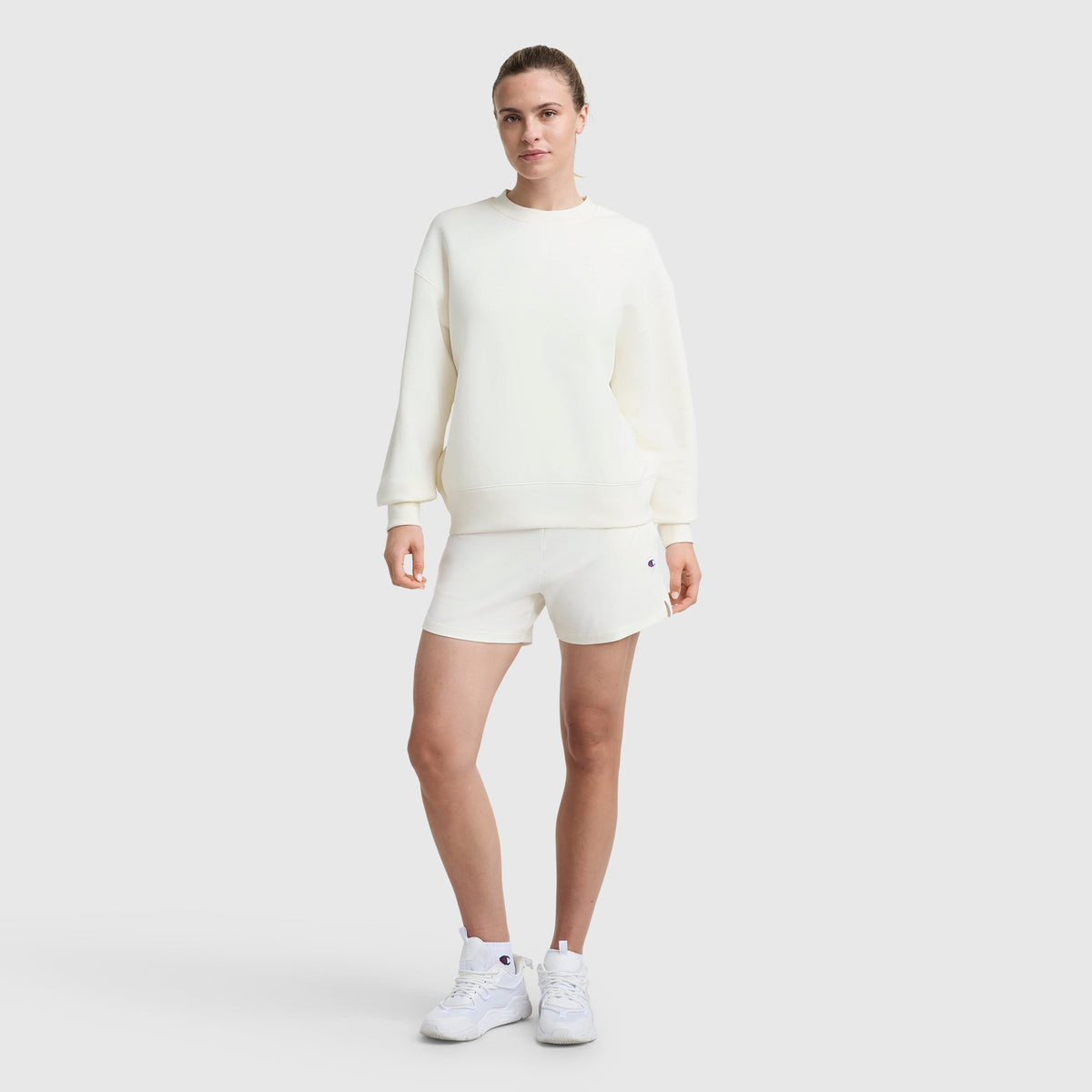 A person stands against a plain light gray background wearing a Champion Powerblend Crewneck Small "C" midweight fleece sweatshirt, paired with matching white shorts and sneakers, one hand by their side and the other slightly bent.