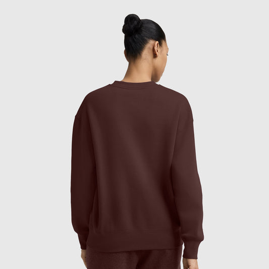 A person with dark hair in a bun is shown from the back, wearing a loose maroon Champion Powerblend Crewneck Sweatshirt with an embroidered script logo and matching pants, standing against a plain light gray background.