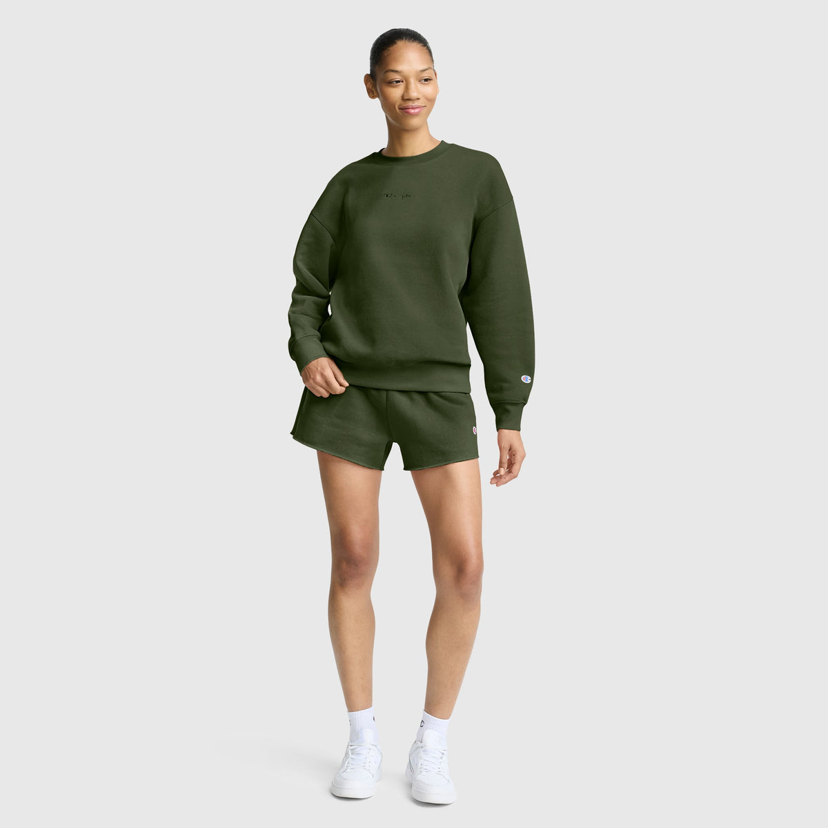 A woman stands against a plain background wearing a Champion Powerblend Crewneck Sweatshirt with an embroidered script logo in olive green, paired with matching shorts and white sneakers. Smiling with one hand in her pocket, she effortlessly showcases the latest in women’s fashion featuring Champion’s cozy Powerblend fleece.