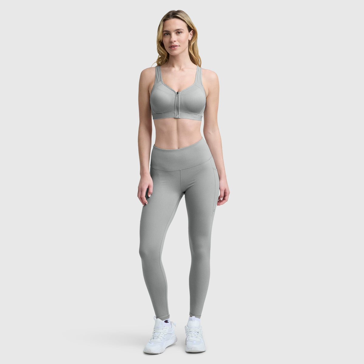 A woman stands against a plain light gray background, wearing the Champion Motion Control Zip Sports Bra, matching high-waisted leggings, and white athletic shoes. She faces forward with a neutral expression.