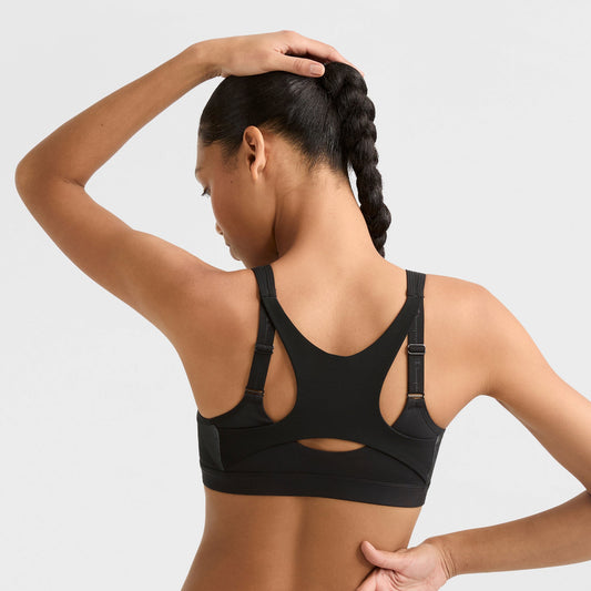 A woman with a long braid is seen from behind, wearing the Champion Motion Control Zip Sports Bra, Wirefree, Script Logo in black, with one hand on the back of her head against a plain light background.