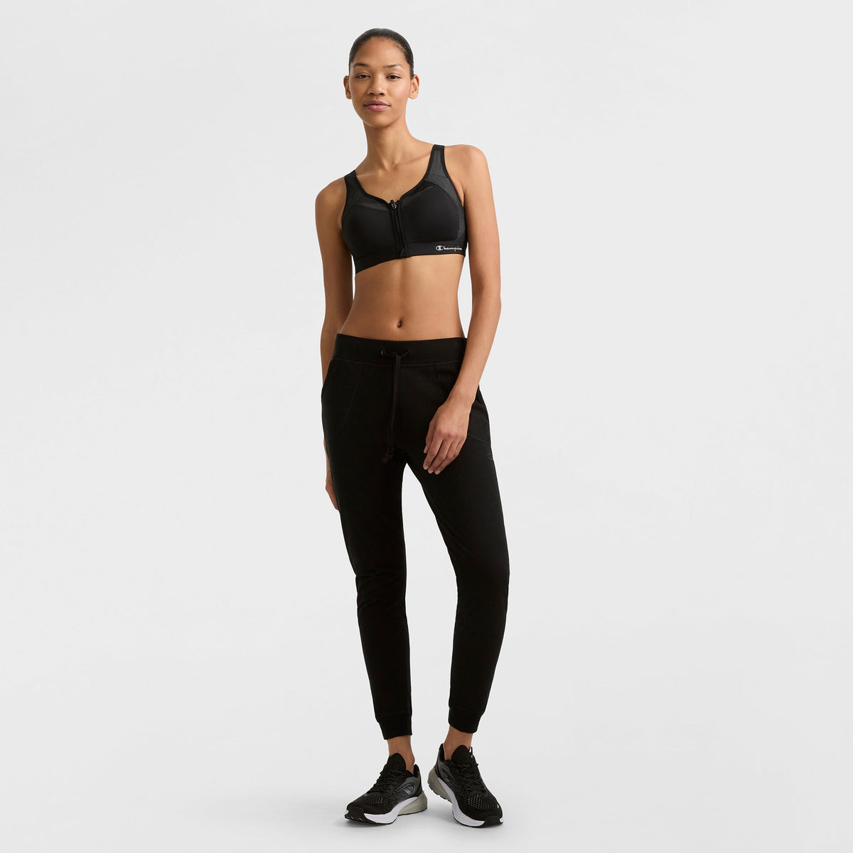 A woman stands against a white background wearing the Champion Motion Control Zip Sports Bra, Wirefree with Script Logo, paired with black jogger pants and black athletic shoes. She looks relaxed, one hand in her pocket and the other by her side.