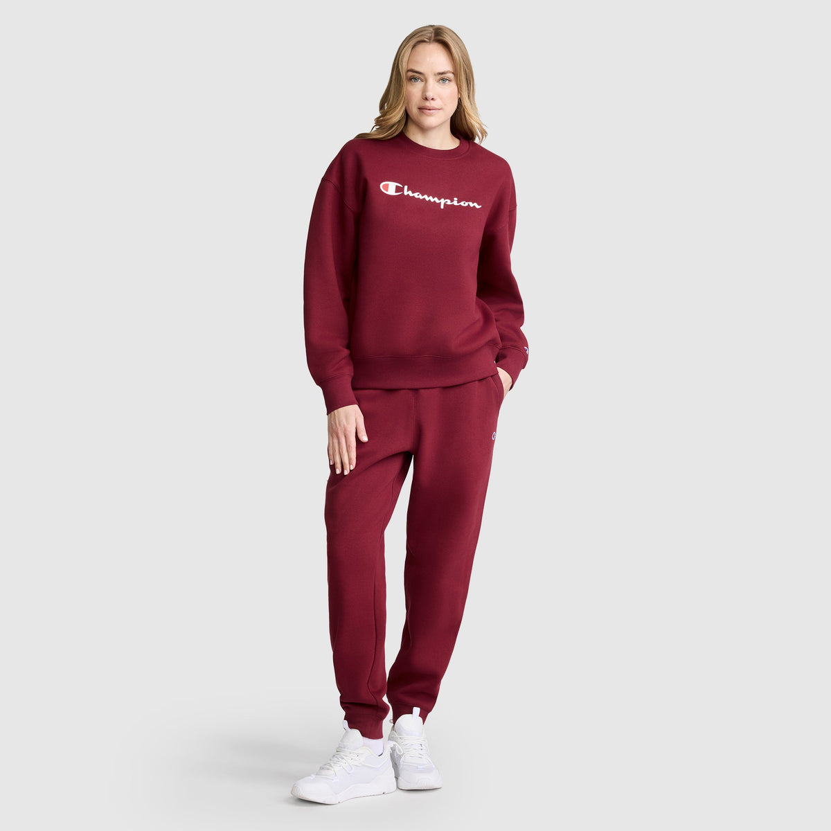 A woman stands wearing the Champion Powerblend Crewneck Sweatshirt with Script Logo in maroon, paired with matching sweatpants and white sneakers. One hand rests in her pocket as she looks at the camera against a plain gray background.