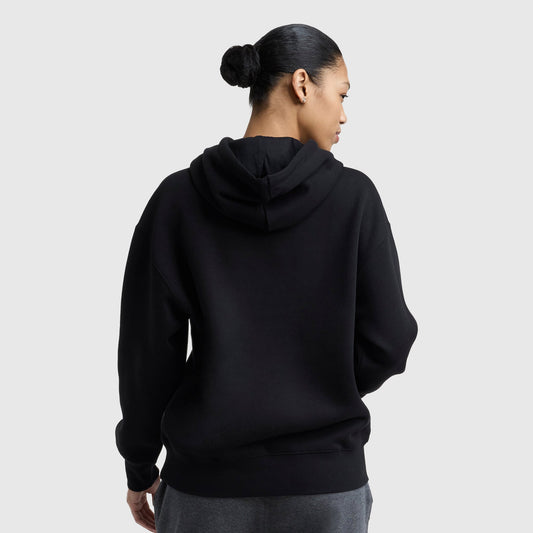 A person with dark hair in a bun is shown from the back wearing a black Champion Powerblend Relaxed Hoodie and gray pants against a plain light background.