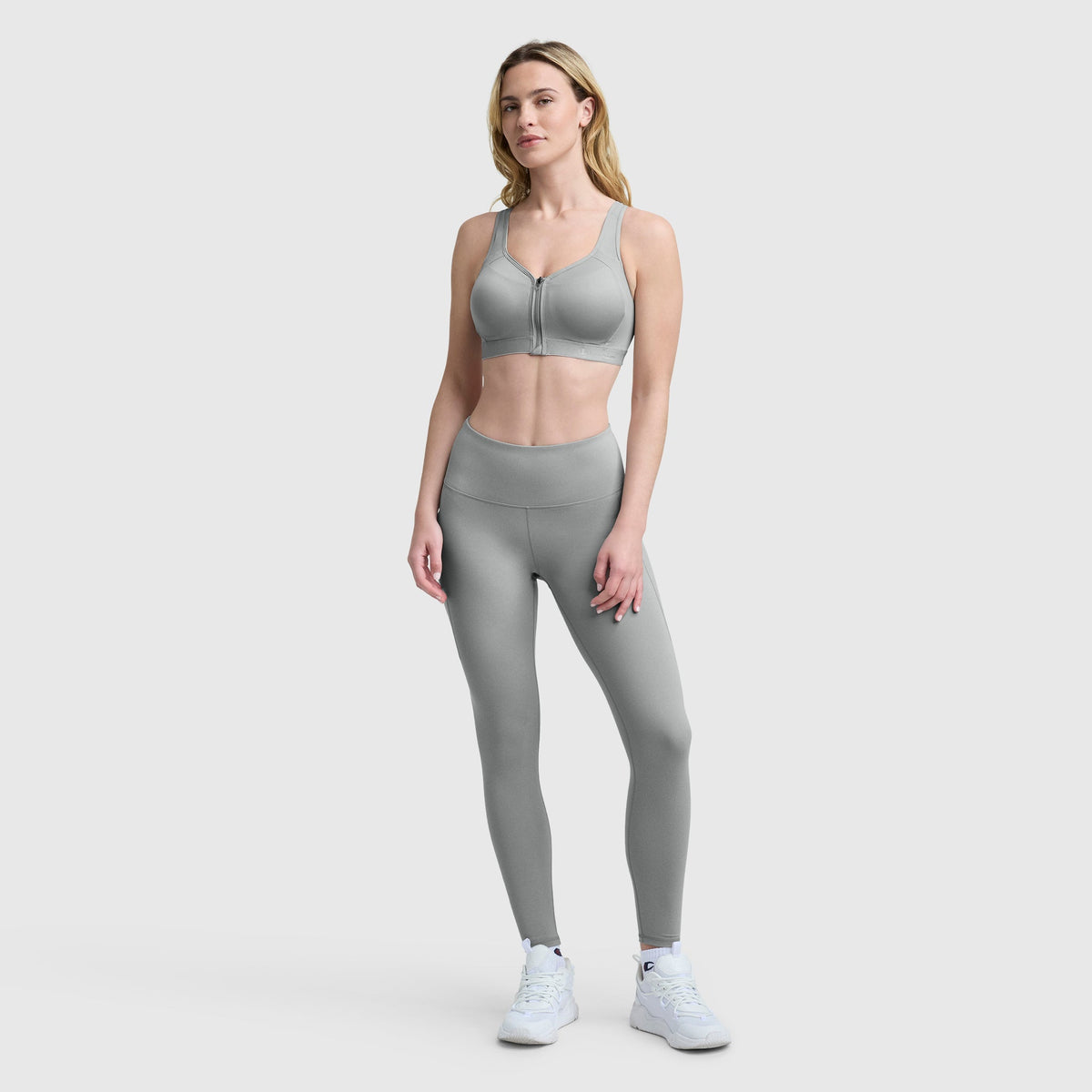 A woman poses facing forward against a light background, wearing Champion Leggings with the C Logo, a light gray sports bra, and white sneakers. One hand rests on her thigh.