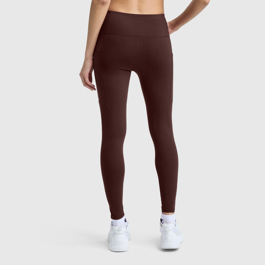 A person wearing Champion C Logo high-waisted, dark brown silky soft leggings and white sneakers stands with their back to the camera against a plain light gray background.