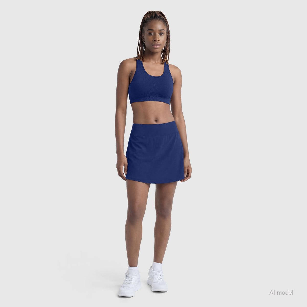 A woman with braided hair stands facing forward in the Champion Courtside Skort (C Logo) and matching sports bra, paired with white sneakers. She poses against a light gray background, showcasing a classic court style look.