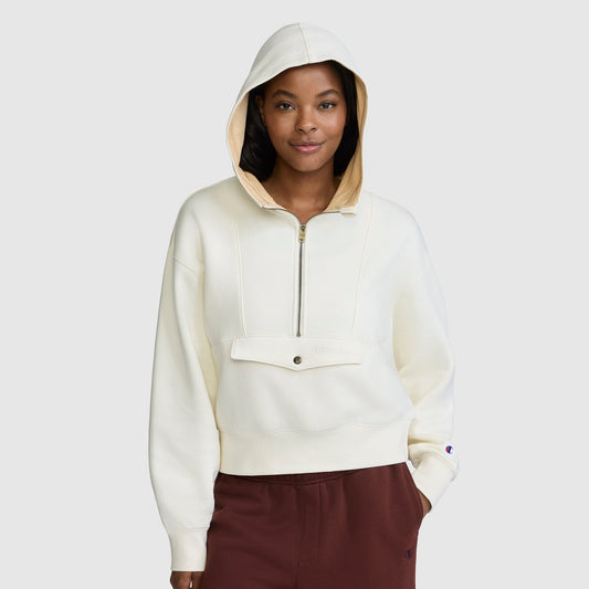 A woman stands against a plain light gray background wearing a Champion Training Half-Zip Fleece Hoodie in cream, which features a front zipper and pocket, paired with maroon pants.