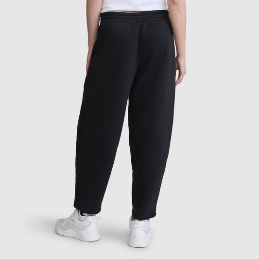 A person stands with their back to the camera against a plain light background, wearing Champion Barrel Leg Fleece Pants made from midweight cotton-poly fleece, paired with a white T-shirt and white sneakers.