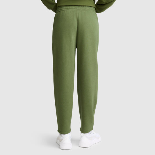A person wearing Champion Barrel Leg Fleece Pants in olive green and white sneakers stands facing away with hands tucked in sweatshirt pockets, set against a plain light gray background.