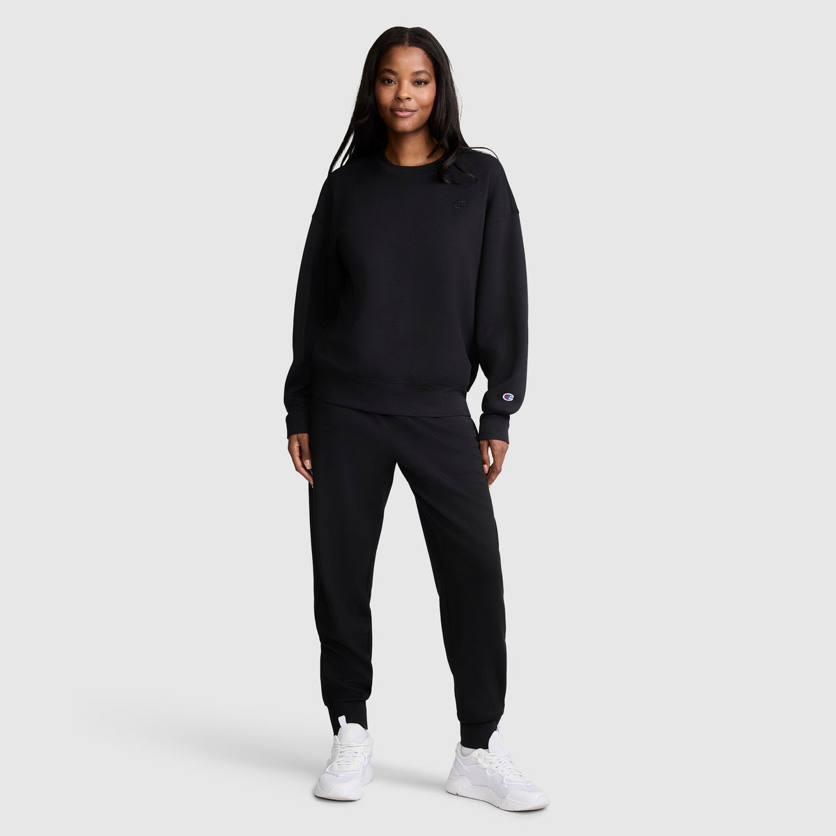 A woman stands smiling, wearing a black Champion Taper Leg Jogger with Zippers and matching joggers paired with white sneakers, against a plain light gray background.