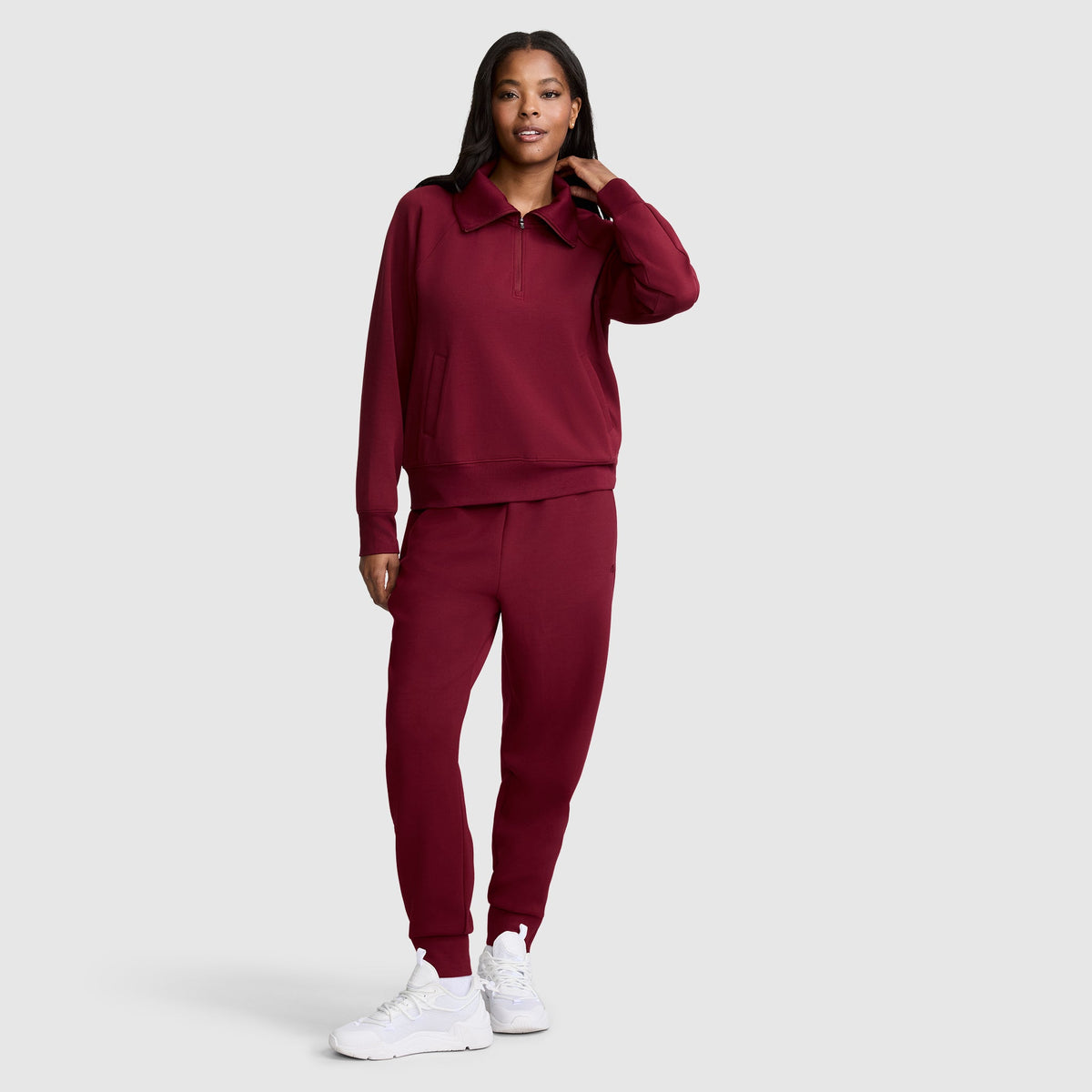 A woman stands against a plain background wearing a matching maroon Champion Taper Leg Jogger with Zippers set and white sneakers. She has one hand touching her hair and smiles slightly.