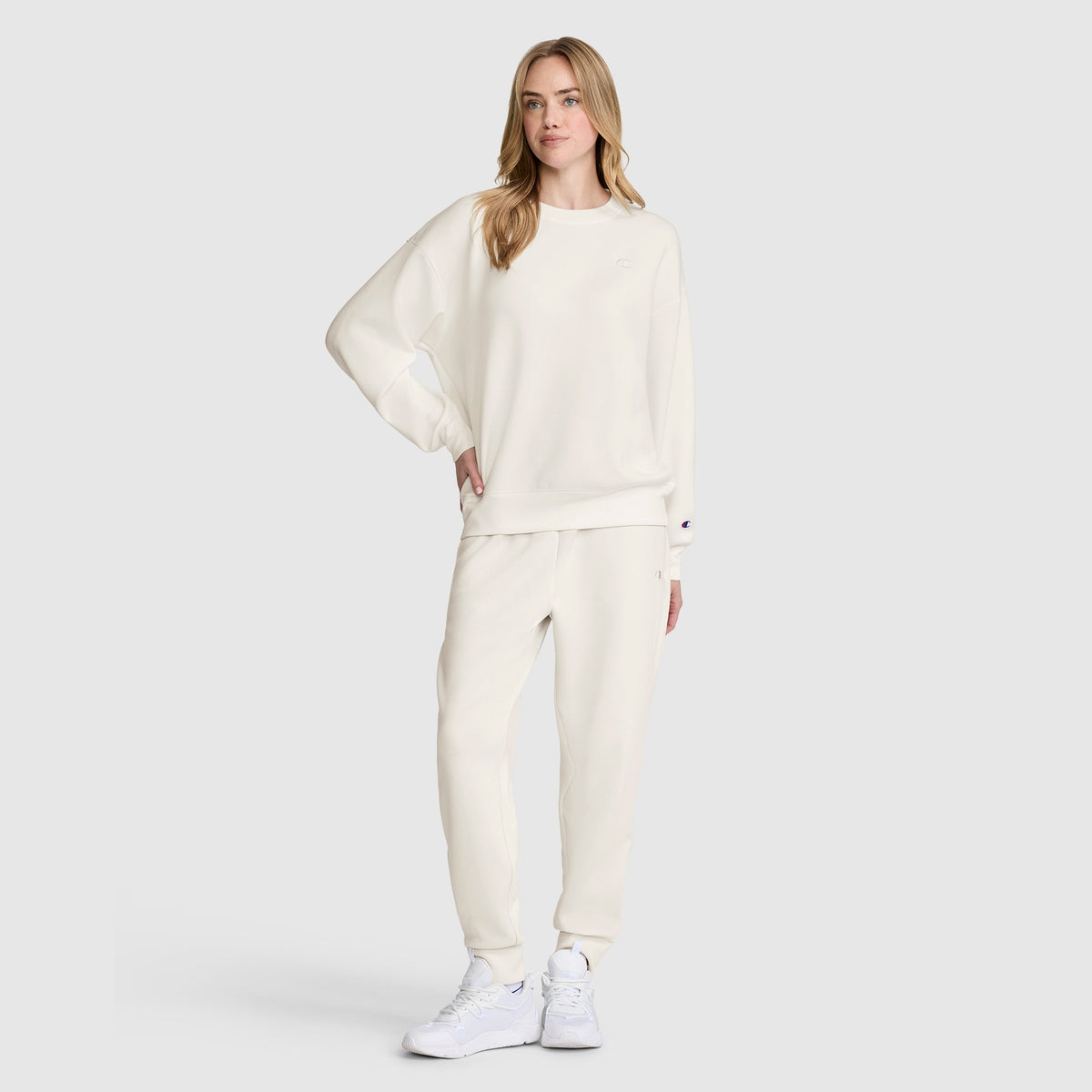 A woman stands against a plain light gray background, wearing an all-white modal scuba blend sweatshirt and Champion Taper Leg Jogger with Zippers, paired with white sneakers.