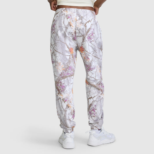 A person wearing white sneakers and Champion’s Powerblend Boyfriend Sweatpant with an allover gray, purple, and orange tree-branch print stands against a plain light background, facing away. The joggers feature a cinch bottom design that gives a modern update to classic Champion C Logo women’s sweatpants.