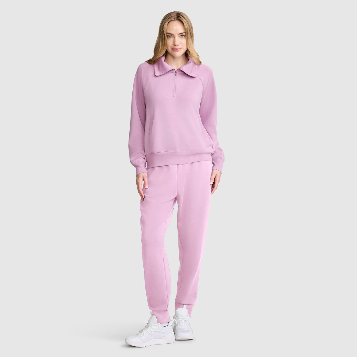 A woman stands facing forward against a plain light gray background, wearing a matching light pink Champion Quarter-Zip Pullover tracksuit and white sneakers. She is smiling softly, with her left hand in her pocket.
